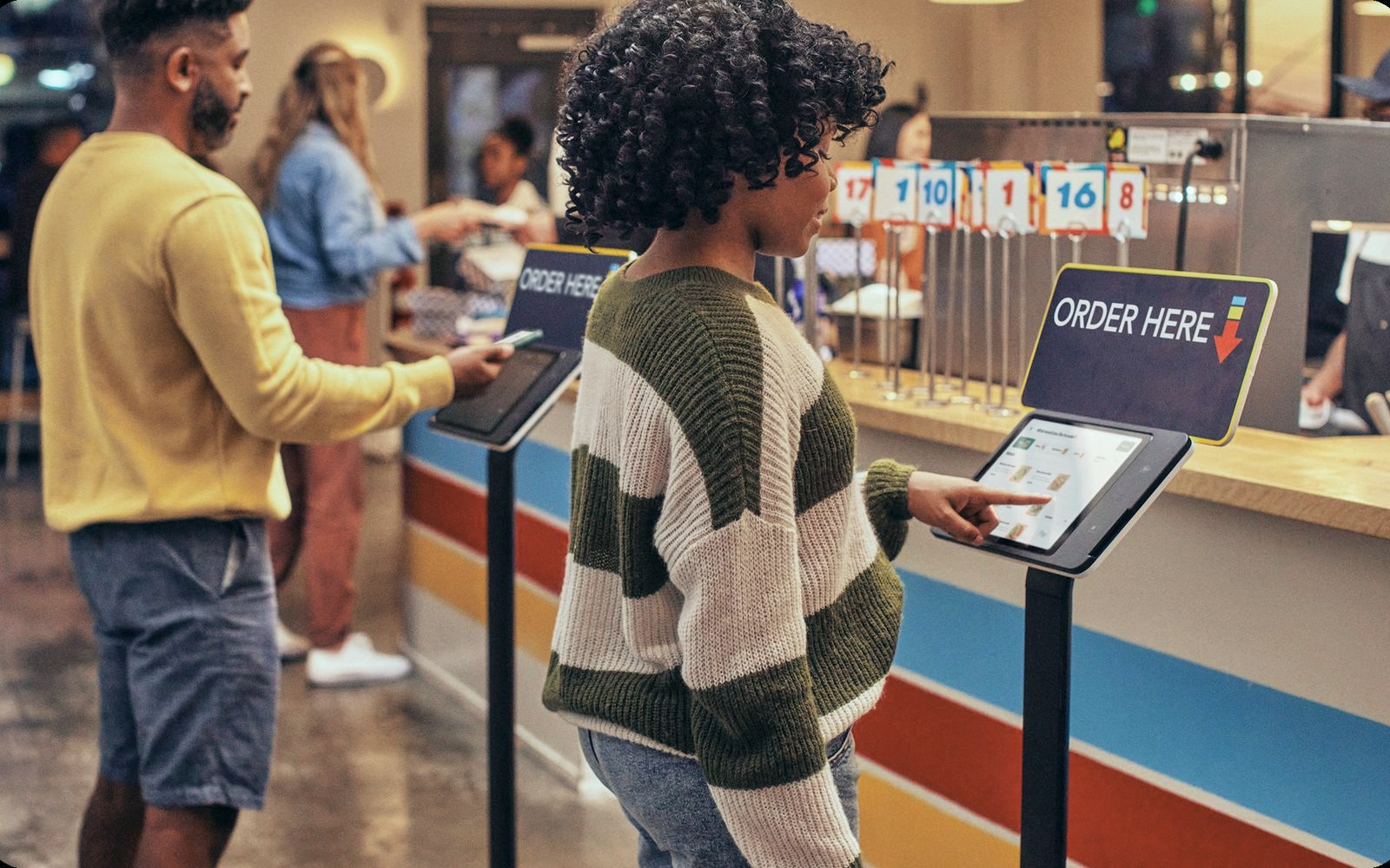 Guests order at two Square Kiosks on floor stands under Order Here signs in a quick-service restaurant